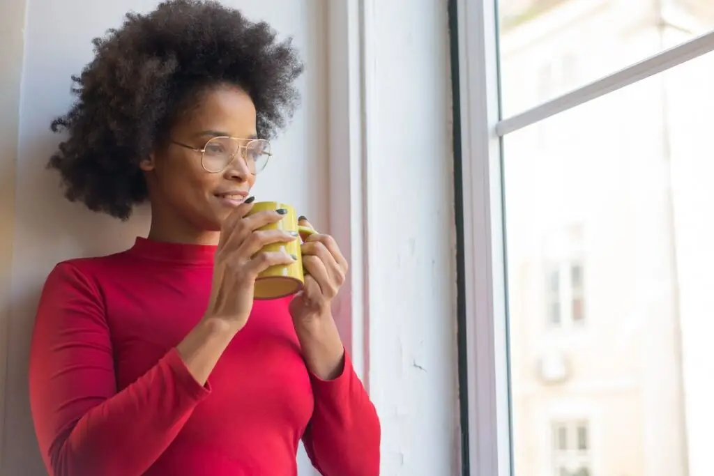 A woman in a red sweater holds a yellow mug and looks out a sunlit window with a calm expression.