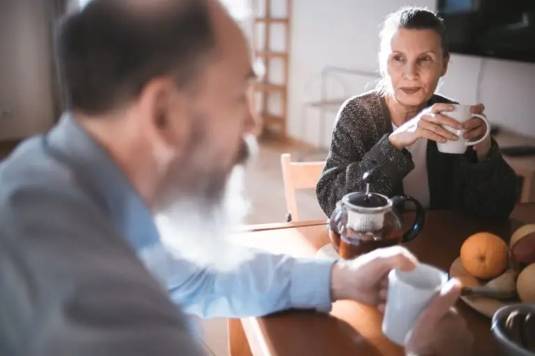 An older woman holding a mug of tea listens to a man across a sunlit kitchen table.