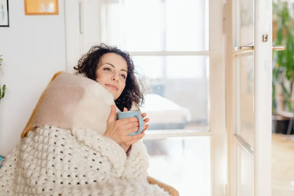A woman wrapped in a knitted blanket sits by a sunny window with a mug of tea, smiling.