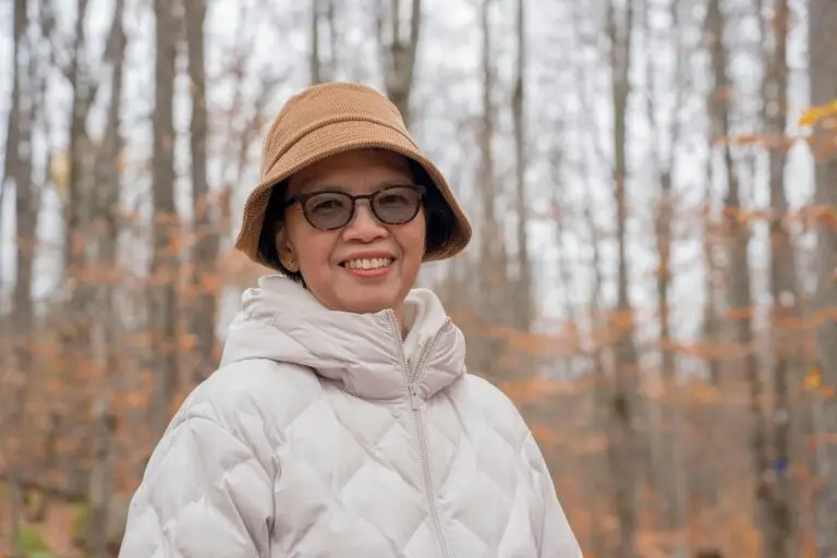 A smiling woman in a bucket hat and glasses stands in an autumn forest.