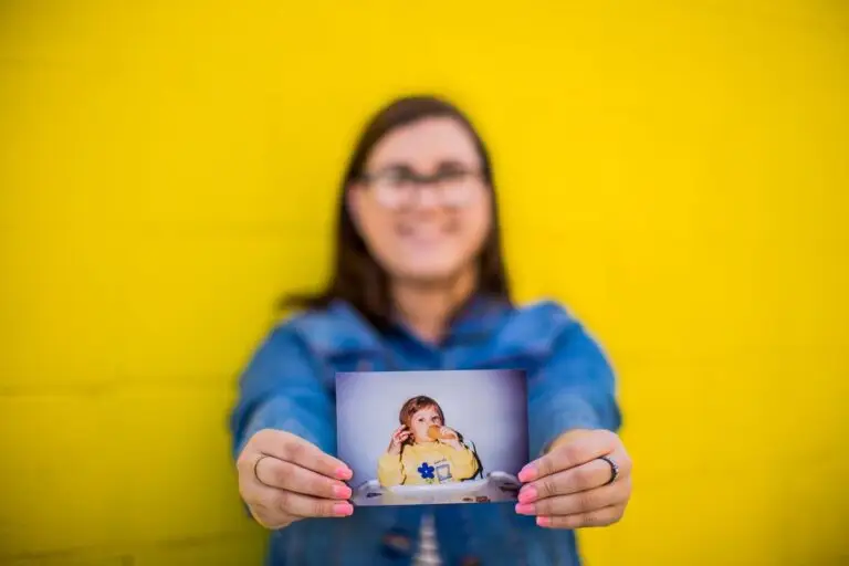 A smiling woman holds up a childhood photo of a toddler in a yellow sweater against a bright yellow wall.