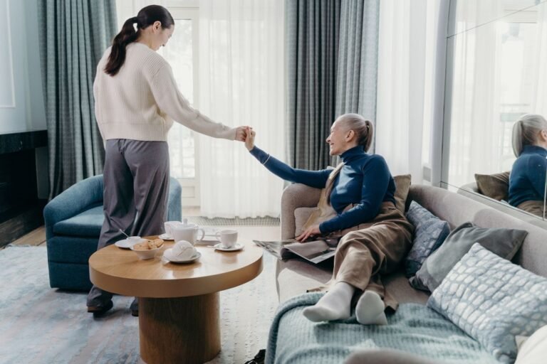 A young woman takes the hand of an older woman resting on a sofa in a bright living room.